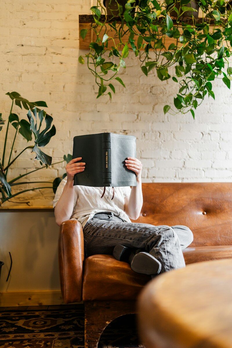 Photo by Humble Lamb A person sitting on a leather sofa reading a black book in a room